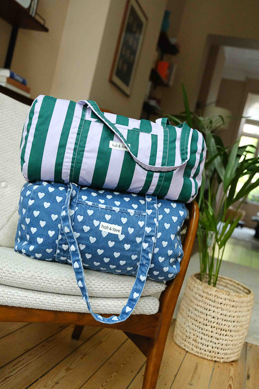 Sac weekend en denim motif cœur et sac weekend stripes, rayures vert et violet, posés l'un sur l'autre sur une chaise. Sac de voyage et accessoire de mode pour femme en coton.