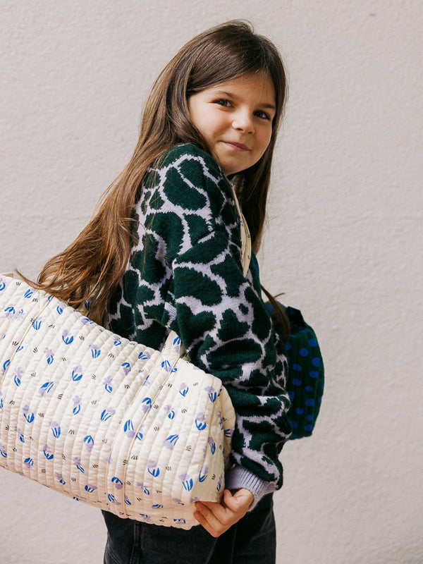 Une adorable petite fille porte un sac week-end imprimé petites fleurs lilas sur son épaule. Ce sac élégant et pratique est idéal pour les mamans et les enfants. Fabriqué en coton bio, il est un accessoire de mode éthique et intemporel.