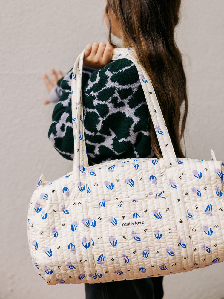 Une adorable petite fille porte un sac week-end imprimé petites fleurs lilas sur son épaule. Ce sac élégant et pratique est idéal pour les mamans et les enfants. Fabriqué en coton bio, il est un accessoire de mode éthique et intemporel.