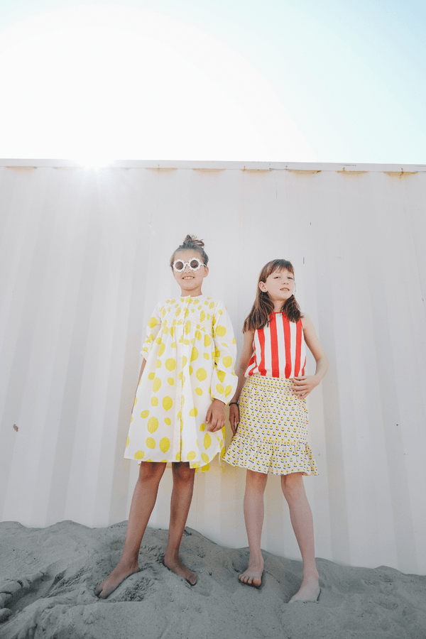 Deux filles jouant sur la plage, l'une portant la robe EDNA motif citron et l'autre le débardeur MARCEL à rayures rouges et la jupe IRMA imprimé petites fleurs jaunes, vêtements en coton bio certifié GOTS style vintage mixte unisexe