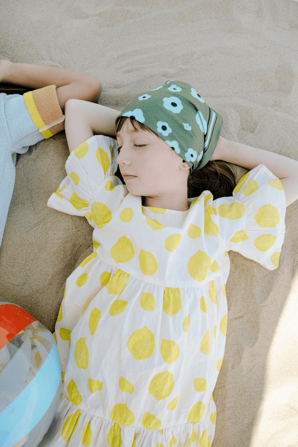 Jeune fille se reposant sur la sable, portant la robe EDNA imprimé citrons et le foulard BRIGITTE à motif grandes fleurs vertes, en coton 100% bio style vintage mixte unisexe