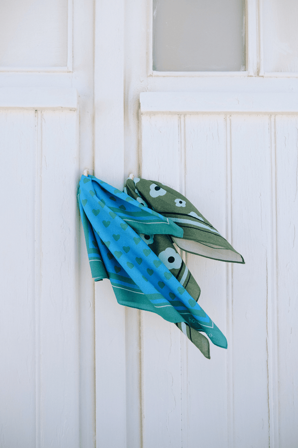Foulards BRIGITTE imprimés bleu avec cœurs verts et grandes fleurs vertes accrochés à une porte de cabine de plage, accessoires en coton bio style vintage mixte